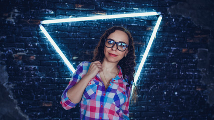 Young woman with glasses in colored plaid shirt posing on background shabby brick wall with glowing...