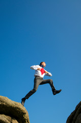 Serious superhero businessman jumping across a chasm between two boulders outdoors against bright blue sky