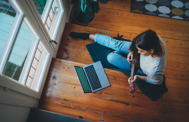 Beginner Woman playing ukulele watching a video tutorial using laptop computer. Young beautiful Woman in casual clothes playing ukulele guitar sitting on the wooden stairs at home, Learning Concept