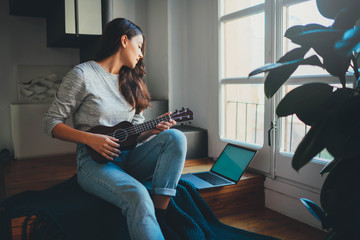 Learning Concept, Young beautiful woman beginner watching video lesson on laptop computer how to play ukulele guitar sitting at wooden stairs at modern home interior with big window, Leisure Time - Powered by Adobe
