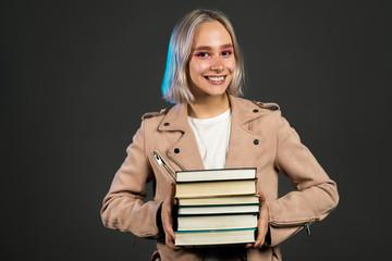 Pretty female student on grey background in studio holds stack of university books from library. Woman smiles, she is happy to graduate.