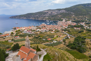 Naklejka na ściany i meble Aerial panoramic view of the cathedral St.Nicholas in Komiza city - the one of numerous port towns in Croatia, orange roofs of houses, picturisque bay, mountain is on background