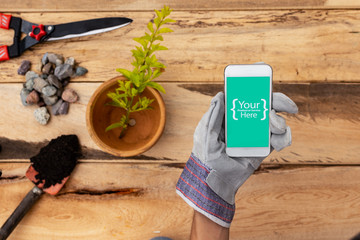 Hand holding smart phone accompanied by garden scissors, flower pot, hand shovel with black soil, gravel on wooden background. Digital or technology services for gardening and landscaping.