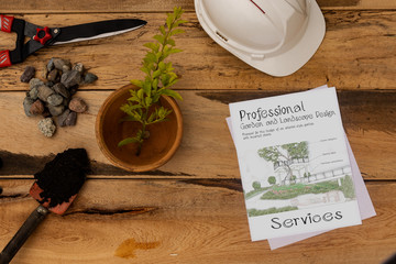 White engineering helmet, sketch garden design, garden scissors, flower pot, hand shovel with black soil and gravel on wooden background. Topical garden and home.