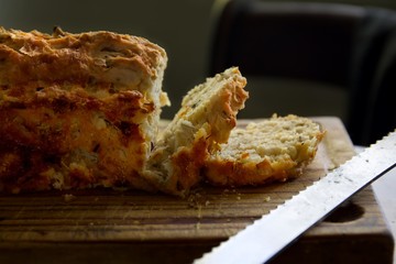 Slices of crusty onion, garlic and cheese beer bread loaf on a wooden board and bread knife to the side.  