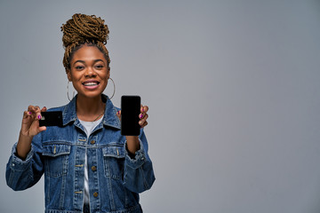 A girl with a smile with hair style in a jeans jacket holds smartphone in one hand and shows a...