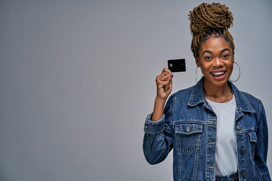 Happy lady with bun in a jeans jacket shows a black bank credit card in her hand. Banking concept