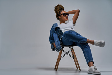 Confident lady in sunglasses in white t-shirt in jeans with denim jacket posing on camera sitting on chair with leg lies on the knee. Fashion concept