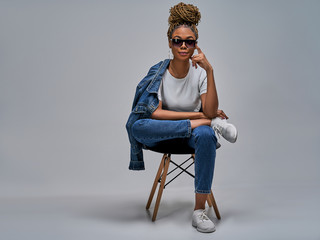 Confident girl in sunglasses in white t-shirt in jeans in sneakers posing on camera sitting on...