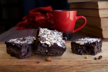 Close up view of three chocolate brownies on a wooden board in front of a red mug and a stack of books in the background.  The brownies have nuts and castor sugar on the top
