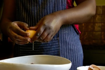 Cook breaking an egg into a mixing bowl.  Black female with blue and white striped apron partially...