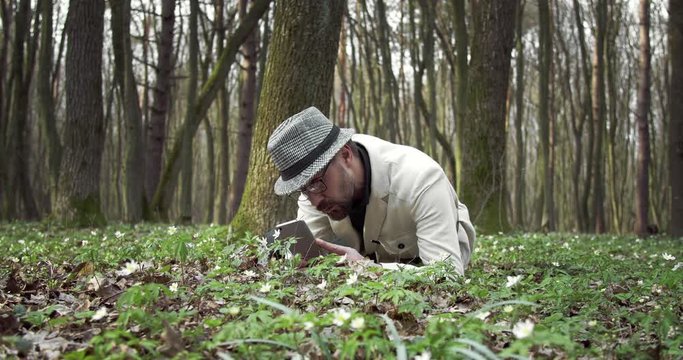 Confident botanist in white jacket and hat lying on ground at forest and taking photo of green plants on smartphone. Bearded man examining beautiful nature around.