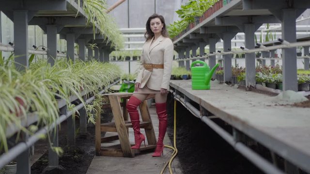 Brunette Caucasian woman in elegant jacket and red thigh high boots standing in glasshouse. Gorgeous lady posing in greenhouse. Nature, lifestyle, beauty.