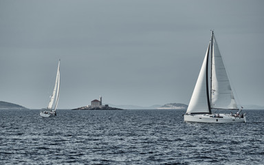 Fototapeta premium Sailboats compete in a sailing regatta at sunset, sailing race, reflection of sails on water, multi-colored spinaker, boat number aft boats, big white clouds, the island with a lighthouse