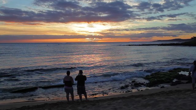 Couple standing at the water's edge at a Poipu beach on Kauai watching a spectacular Hawaiian sunset as gentle waves roll to the shore.