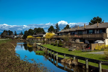 Residential area in a picturesque place on the banks of the canal with thickets of blackberry bushes on the other shore against the spring forest, mountain range and cloudy sky
