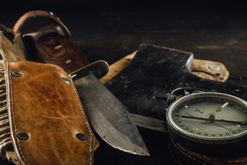 Old military compass, rusty hunting bushcraft knife, and small axe on the dark wooden table....