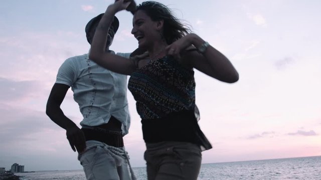 Young man and woman enjoying salsa dance on retaining wall at sunset, sea in background