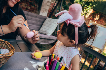 little girl with Easter themed hat and her mother coloring the eggs for Easter decorations in the garden.