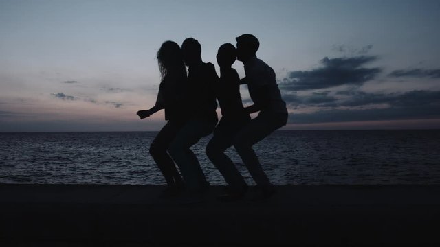 Men and women dancing and having fun on retaining wall at sunset, sea in background