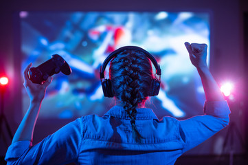 Girl in headphones plays a video game on the big TV screen. Gamer with a joystick. Online gaming with friends, win, prize. Fun entertainment. Teens play adventure games. Back view. Neon lighting - obrazy, fototapety, plakaty