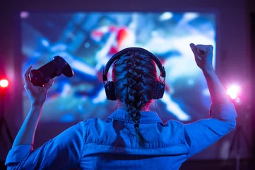 Fotobehang Gaming Girl in headphones plays a video game on the big TV screen. Gamer with a joystick. Online gaming with friends, win, prize. Fun entertainment. Teens play adventure games. Back view. Neon lighting  © Anton