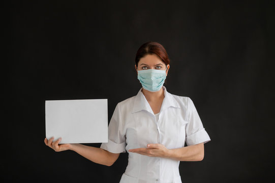 A Young Doctor In A Medical Mask Holds A White Poster On A Black Background. Female Nurse In Uniform Pointing To A Blank Billboard.