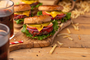 Juicy Burger on a wooden table with a drink. Homemade cheeseburger on the cutting board with beef, cheese, onions, tomatoes, cucumbers, sauce, french fries and sweet soda. Fast food