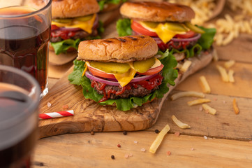 Burger on a wooden table with a drink. Homemade cheeseburger on the cutting board with beef, cheese, onions, tomatoes, cucumbers, sauce, french fries and sweet soda. Food concept.