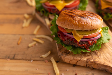 Hot juicy cheeseburger on a wooden cutting board. Burger with beef, cheese, vegetables, crispy buns and salt fries on a rustic table. Junk food concept. Copy space