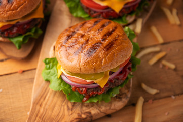 Fresh hot juicy burger with salty french fries. Meal on a cutting kitchen board on a wooden table. Cheeseburger with meat, cheese, tomatoes, cucumbers and onions. Street junk food concept. Top view.