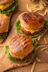 Fresh hot juicy burger with french fries. Meal on a cutting kitchen board on a wooden table. Cheeseburger with meat, cheese, tomatoes, cucumbers and onions. Junk food. Top view. American cuisine