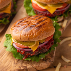 Fresh hot juicy burger with salty french fries. Meal on a cutting kitchen board on a wooden table. Cheeseburger with meat, cheese, tomatoes, cucumbers and onions. Square picture. American cuisine