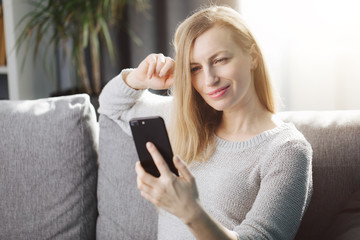 Happy mature woman with blond hair sitting on grey couch and looking at smartphone screen. Smiling female in domestic outfit using phone while relaxing at home.