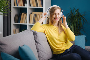 Happy mature woman in casual outfit sitting on couch with closed eyes and enjoying favorite songs in headphones. Smiling lady in yellow sweater relaxing at home.