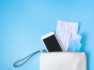 Flat lay of white leather woman bag with smartphone, surgical mask and alcohol gel sanitizer on blue background, Top view with copy space for text. Covid-19 and coronavirus concept.
