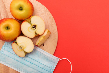 Whole and cut into pieces apples and protective mask lie on a round cutting board on a red background.Harvest coronavirus concept.The concept of food shortage due to the covid-19.