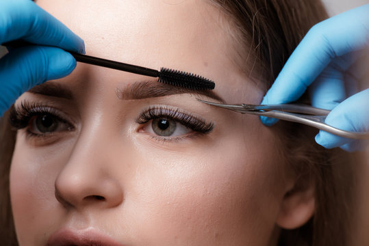 FeMale professional hairdresser serving client, shearing eyebrows with scissors. Ginger stylish young man with thick big beard and short hair getting trendy haircut in black cape. Light barber shop