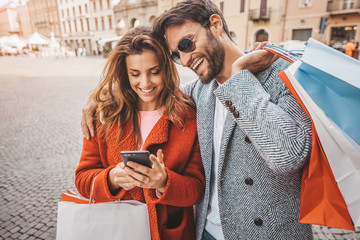 Young beautiful couple doing shopping with smartphone online holding shopping bags in the city. 