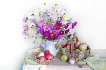 Still life with a bouquet of pink flowers in a vase and a basket of natural, ripe apples on a light background