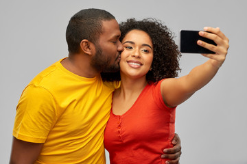 technology and people concept - happy smiling african american couple taking selfie by smartphone and kiss over grey background