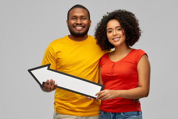direction and people concept - happy smiling african american couple holding big leftwards thick arrow over grey background