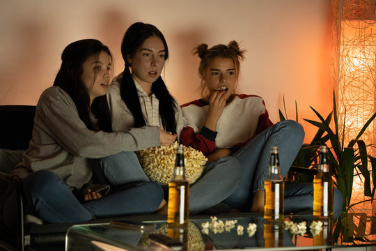 Three friends watching movie on tv, sitting on a couch at home and eating popcorn. Three beautiful girls watching scary film and laughing. Stay at home concept. 