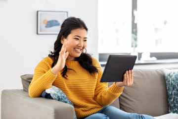 people and leisure concept - happy smiling asian young woman in yellow sweater with tablet pc computer having video call and waving hand at home