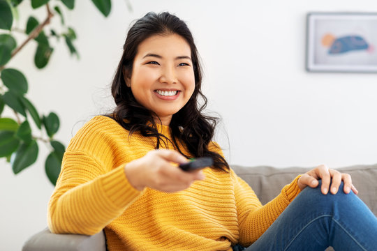 People And Leisure Concept - Happy Smiling Asian Young Woman With Tv Remote Control Sitting On Sofa At Home