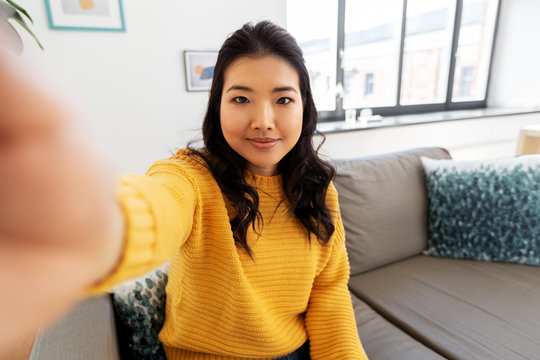 people and leisure concept - happy smiling asian young woman in yellow sweater taking selfie at home