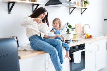 happy mom and daughter are drinking tea sitting on the table in the kitchen. white teapot, porcelain Cup in his hands. mom brunette in white shirt and jeans, daughter in denim overalls.
