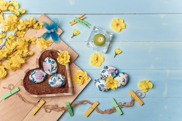 Heart shaped box with decorated Easter eggs, yellow flowers, burning candle and brown boards on wooden blue background. Spring and Easter concept. Top view, flatlay, copy space.