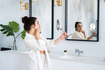 Young beautiful woman is standing in the bathroom, putting on lipstick and looking in the mirror. Sexy girl dressed in white shirt, pants and bra. Beauty has dark hair and eyes, plump lips.