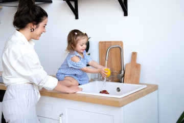 A beautiful young woman with her two year old daughter is washing fruit in the kitchen sink. Fresh bright fruits, apples, oranges, pears. Healthy food for children. The baby helps her mother.